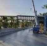 Blue crane beside an unfinished elevated pedestrian bridge over a roadway, with guardrails and trees nearby.