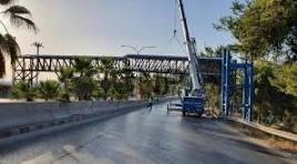 sal - Center News Network Blue crane beside an unfinished elevated pedestrian bridge over a roadway, with guardrails and trees nearby.