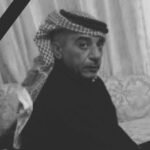 Black-and-white portrait of a man wearing a keffiyeh headscarf and dark clothing, looking toward the camera with a serious expression, mourning ribbon in the corner.