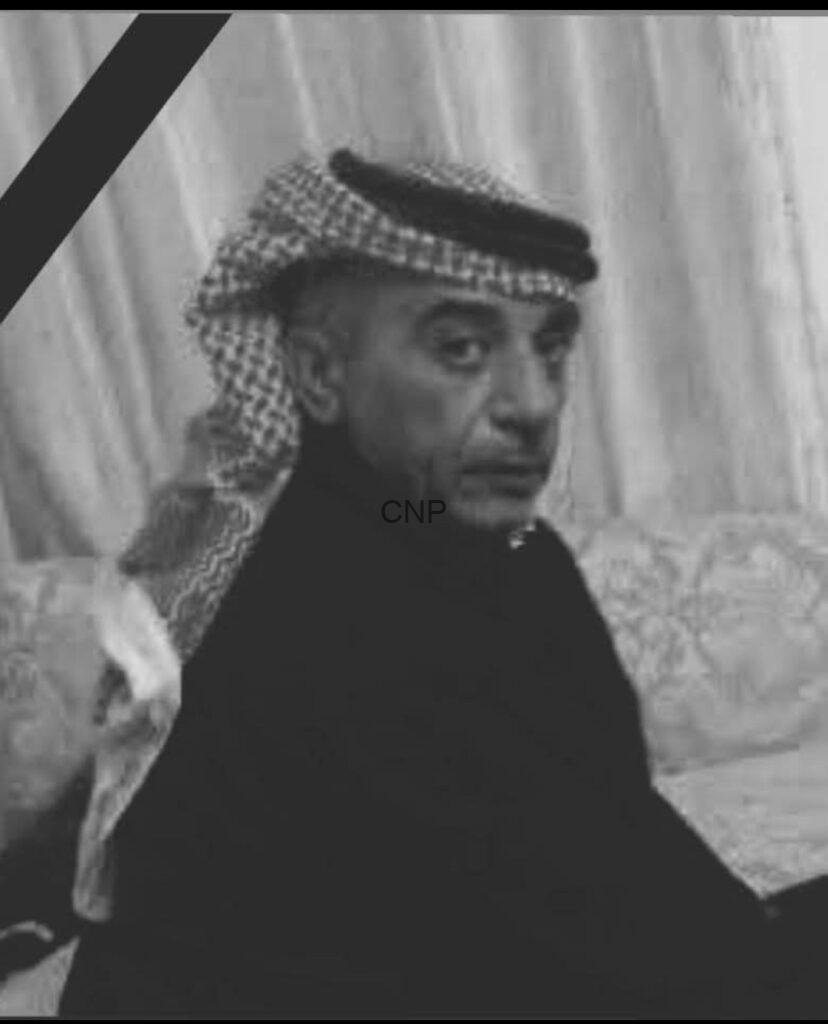 Grayscale photo of a man wearing a keffiyeh and dark robe, looking to the side, with a diagonal black mourning ribbon across the corner.