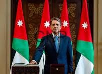 Man in a blue suit speaks at a podium with Jordan flags behind him in a formal government setting.【no punctuation】],