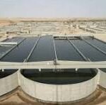 Rooftop solar panels on a low industrial building in a desert, with circular vent structures in the foreground and a blue sky above.