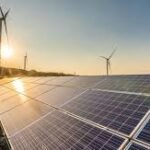 Solar panels in a field with wind turbines on the horizon at sunset, illustrating renewable energy.
