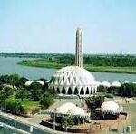 White-domed mosque complex with a tall central minaret beside a river and surrounding greenery.