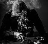 A person in a suit sits at a table in a dark, smoky scene with a skull in front of them.