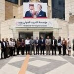 Group of about 25 people posing in front of a beige institutional building with an Arabic banner and a large poster of two men above the entrance, in a courtyard area with a patterned ground.