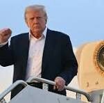 Man with white hair in a dark suit stands on airplane stairs, raising his right fist.