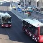 Red tram in the foreground travels along a city street while a white tram and cars pass by, storefronts lining the sidewalk behind.