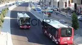 Red tram in the foreground travels along a city street while a white tram and cars pass by, storefronts lining the sidewalk behind.