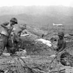 Soldiers digging in a trench with barbed wire in the foreground and a distant airplane in a barren landscape.
