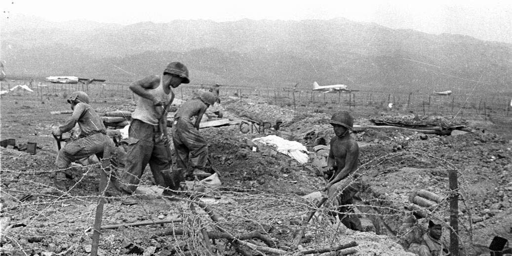 war - Center News Network Soldiers digging in a trench with barbed wire in the foreground and a distant airplane in a barren landscape.