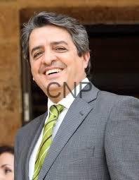 waz - Center News Network Smiling man in a gray suit with a green striped tie, standing outdoors near a brick structure.