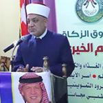 Man in a white traditional head covering speaks at a microphone from a podium at a charity event, with Arabic banners and flags in the background.