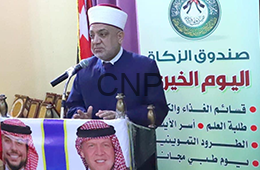 Man in a white traditional head covering speaks at a microphone from a podium at a charity event, with Arabic banners and flags in the background.