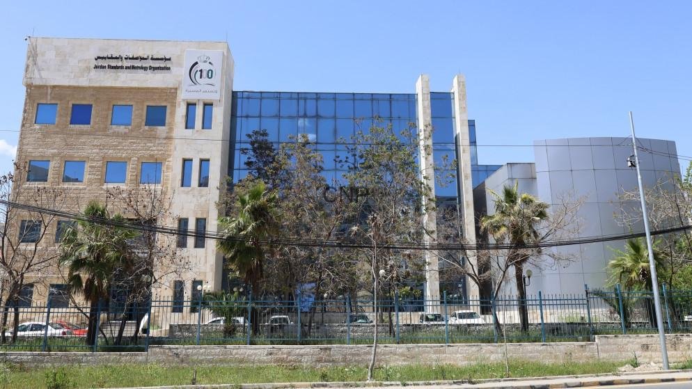Modern office building with a beige stone left wing and blue glass center, fenced and lined with trees under a clear blue sky.