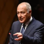 Older man in a dark pinstripe suit delivering a speech at a podium, gesturing with his hands.