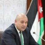 Bald man in a dark suit and green tie seated at a desk with a microphone, flag in the background.