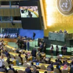 UN General Assembly hall filled with delegates seated in colored rows, large screens display the chamber, and a podium area at front