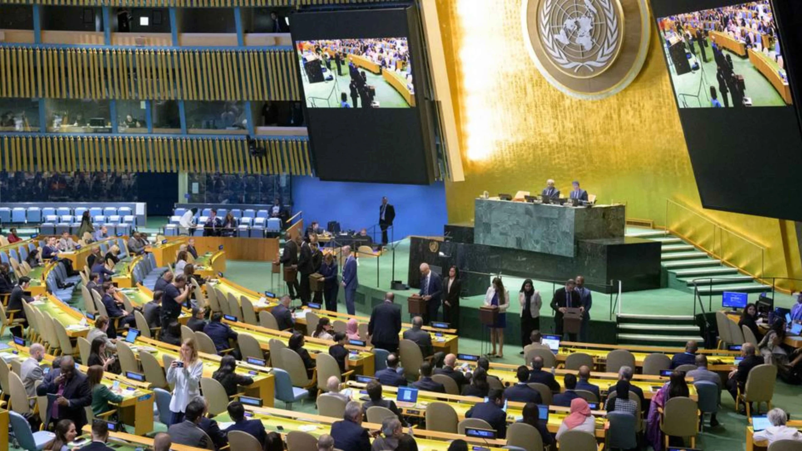 UN General Assembly hall filled with delegates seated in colored rows, large screens display the chamber, and a podium area at front