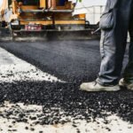 Road construction scene: worker in boots guiding asphalt being laid by a paving machine on a paved surface.