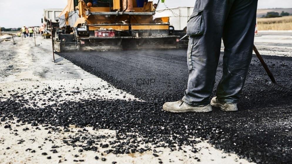 aman - Center News Network Road construction scene: worker in boots guiding asphalt being laid by a paving machine on a paved surface.