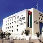 Modern white institutional building with Arabic signage, palm trees, and a Jordan flag on a flagpole in front.
