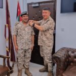 Two soldiers in desert camouflage stand in an office as one pins a medal on the other's chest, with Jordan’s flag and a second flag in the corner and a clock on the wall behind them.
