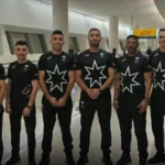 A lineup of athletes in black uniforms with large white star logos, standing in an airport terminal.