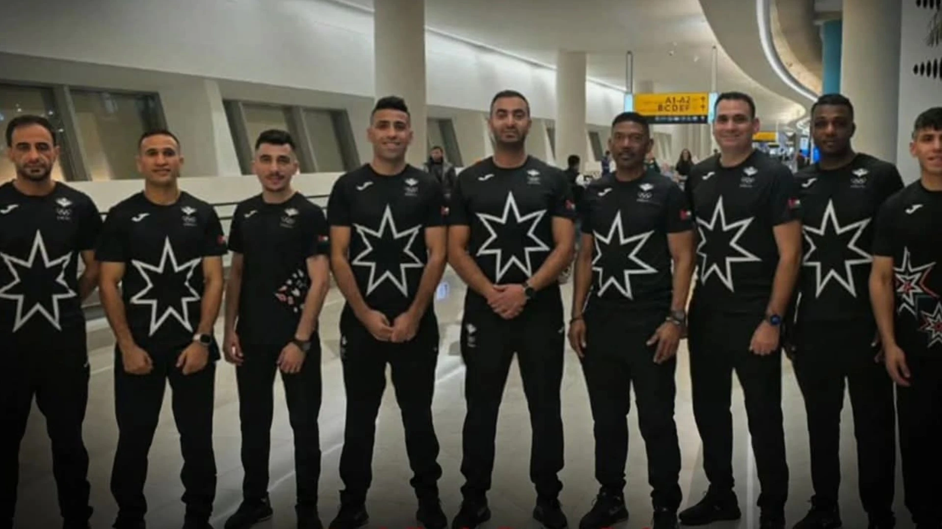 A lineup of athletes in black uniforms with large white star logos, standing in an airport terminal.
