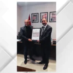 Two men in suits present and receive a framed certificate in an office, with a Jordanian flag and portraits on the wall behind them.