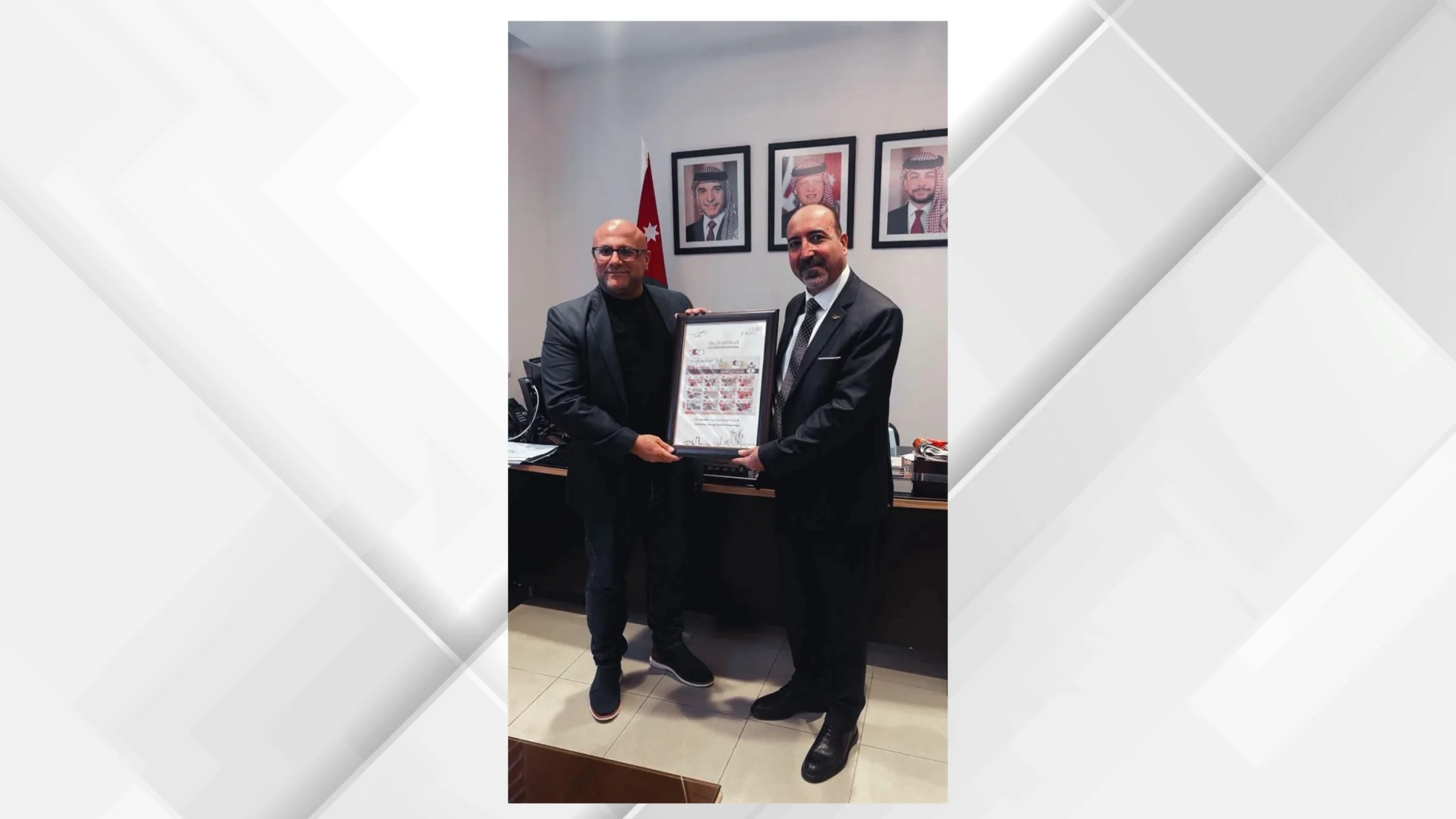 Two men in suits present and receive a framed certificate in an office, with a Jordanian flag and portraits on the wall behind them.