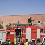 Red fire truck with firefighters on the roof and a ladder extended toward a tan building under a clear blue sky, depicting an emergency response.