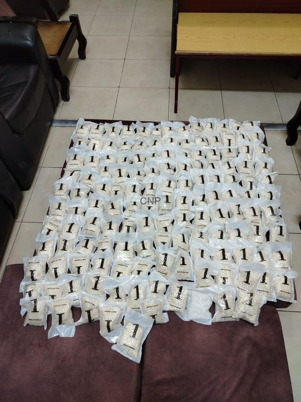 Many small white plastic bags filled with light-colored grains are spread in neat rows on a brown table in a tiled room.