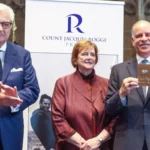 Three people in business attire on a stage; left man claps, center woman smiles, right man holds a plaque in front of a banner reading 'Count Jacques Rogge'.