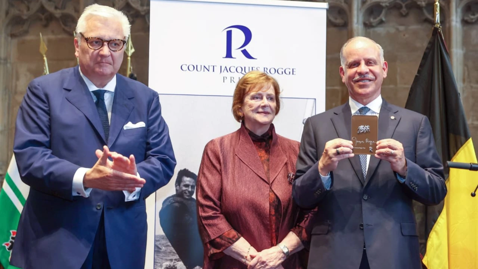 faisal - Center News Network Three people in business attire on a stage; left man claps, center woman smiles, right man holds a plaque in front of a banner reading 'Count Jacques Rogge'.