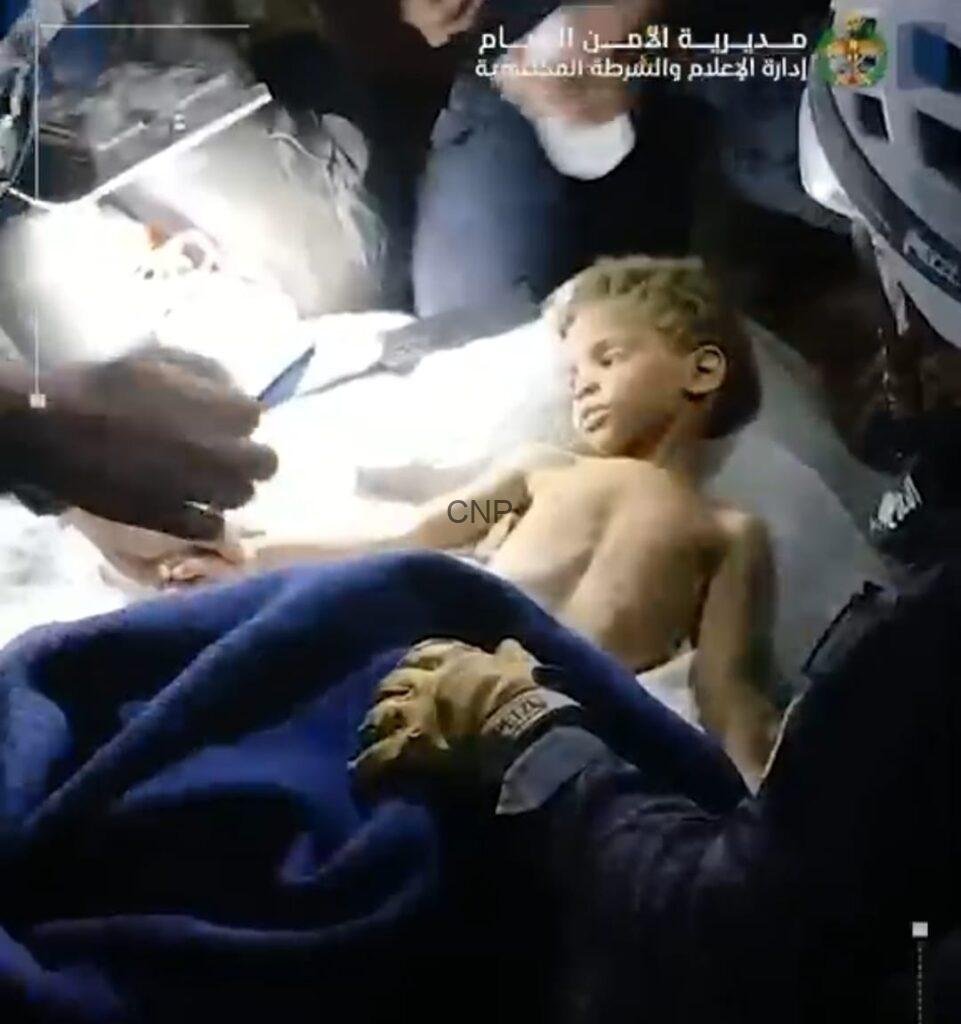 Boy lying shirtless on a hospital bed while medical staff attend to him in an emergency setting, bright light overhead.