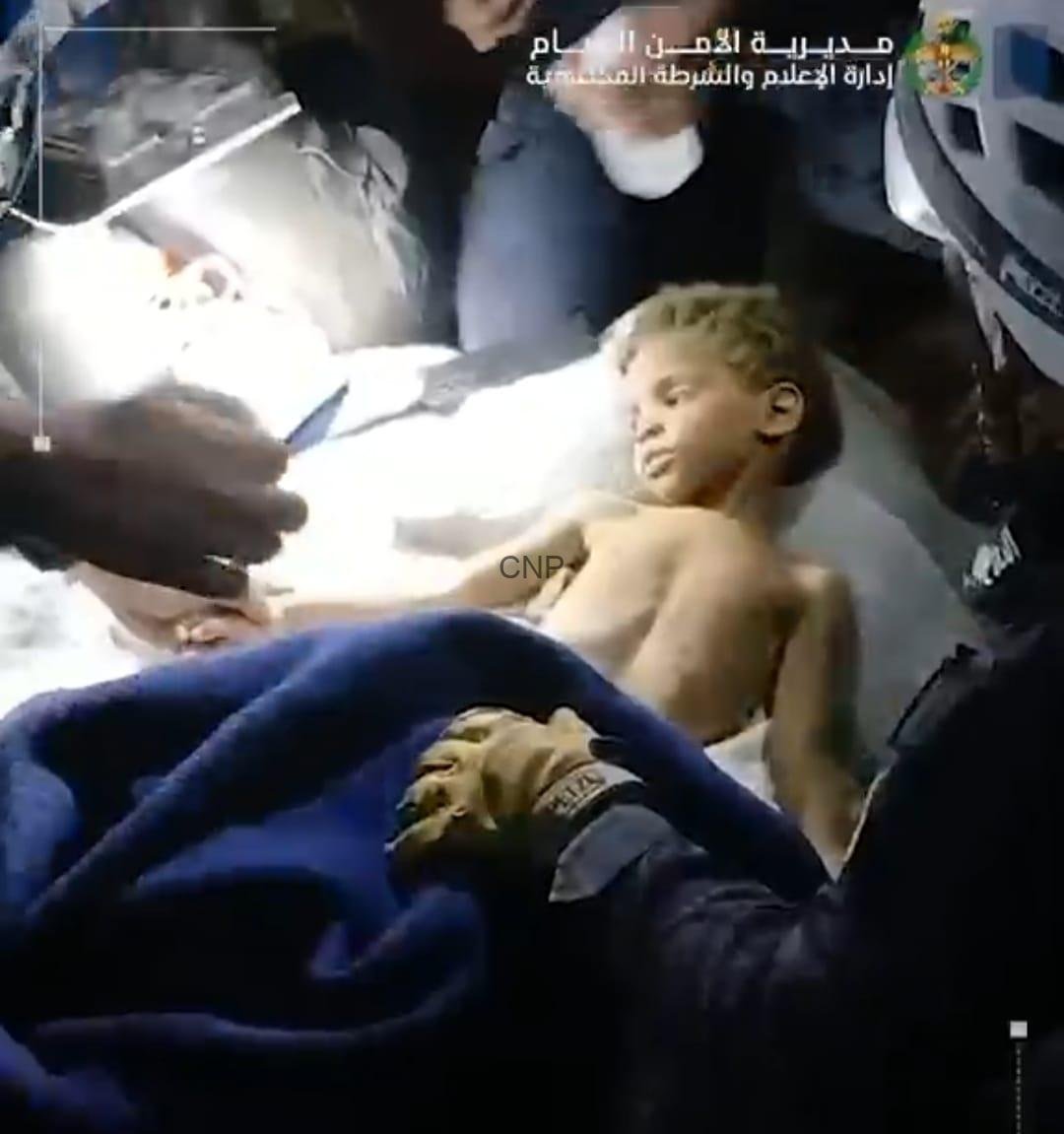 Boy lying shirtless on a hospital bed while medical staff attend to him in an emergency setting, bright light overhead.