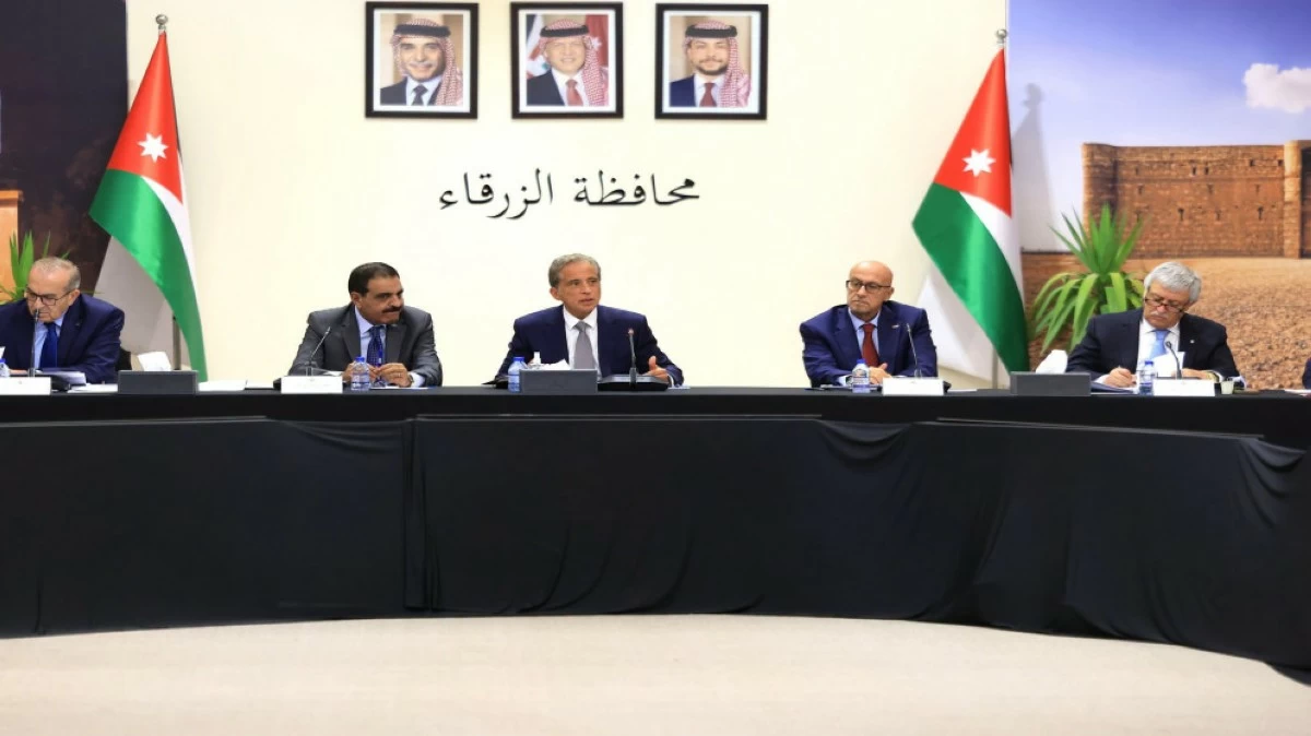Panel of officials seated at a long black-draped table in a formal meeting, Jordanian flags on both sides and microphones in front of each person.