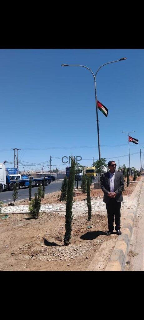 hos2 - Center News Network Man in a suit stands by a landscaped street median with small trees; a flag on a lamppost and trucks are visible in the background on a sunny day.