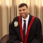 Smiling man in a black graduation gown with red trim and a white shirt, seated indoors against a light curtain backdrop.