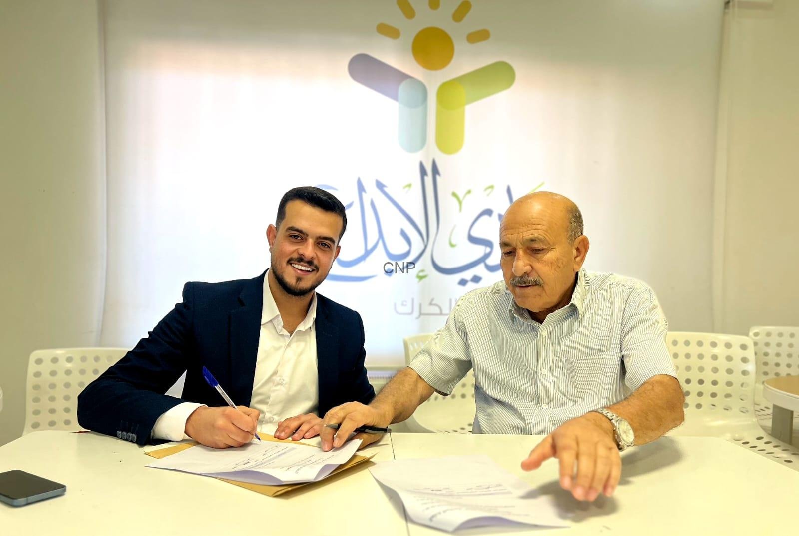 ibdaa - Center News Network Two men sit at a white table signing documents, one in a suit and the other in a striped shirt, with a colorful logo on the wall behind them.