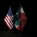 Two flagpoles with the United States and Iran flags on a dark stage, a person in a suit between them holding the Iranian flag.