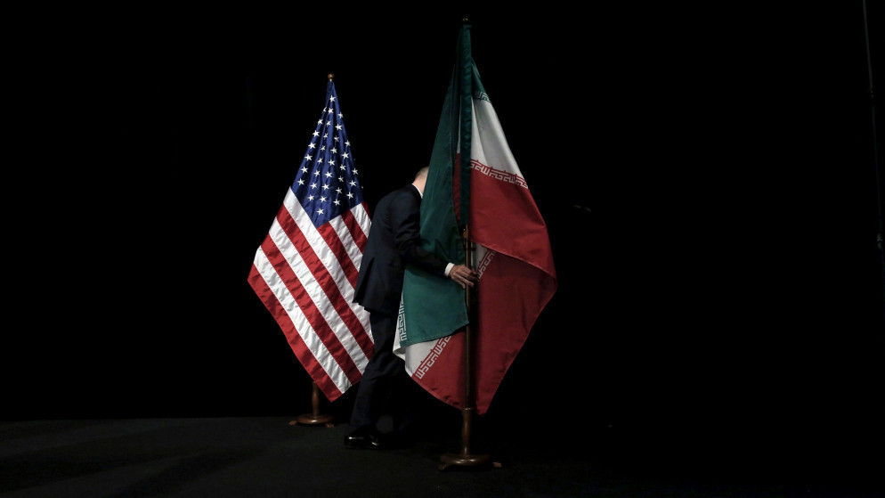 Two flagpoles with the United States and Iran flags on a dark stage, a person in a suit between them holding the Iranian flag.