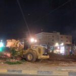 Night street construction site with a large yellow bulldozer moving dirt, storefronts lit in the background, and a couple of people observing nearby.