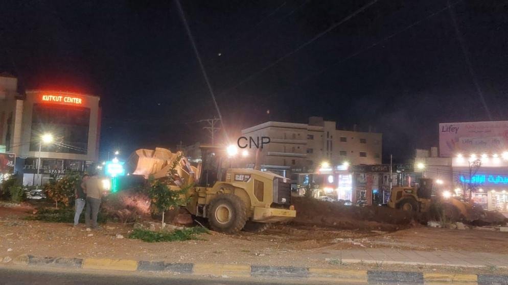 irbed - Center News Network Night street construction site with a large yellow bulldozer moving dirt, storefronts lit in the background, and a couple of people observing nearby.