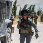 Female soldier in tactical gear running toward the camera, pistol drawn, beside an armored vehicle with teammates in the dusty background.