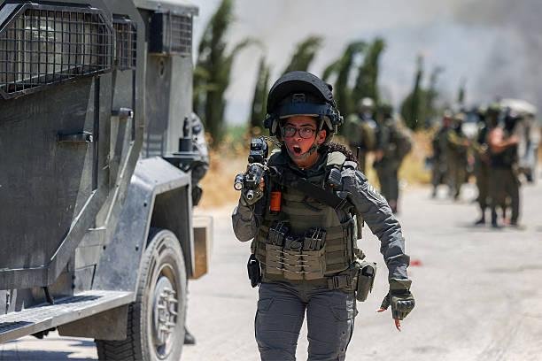 isar - Center News Network Female soldier in tactical gear running toward the camera, pistol drawn, beside an armored vehicle with teammates in the dusty background.