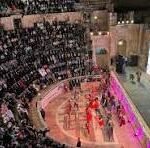 Indoor arena with a circular performance pit and tiered audience seating, performers in red on a brightly lit stage surrounded by spectators.