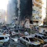 Firefighters amid charred ruins and collapsed cars in a destroyed urban block after a disaster.