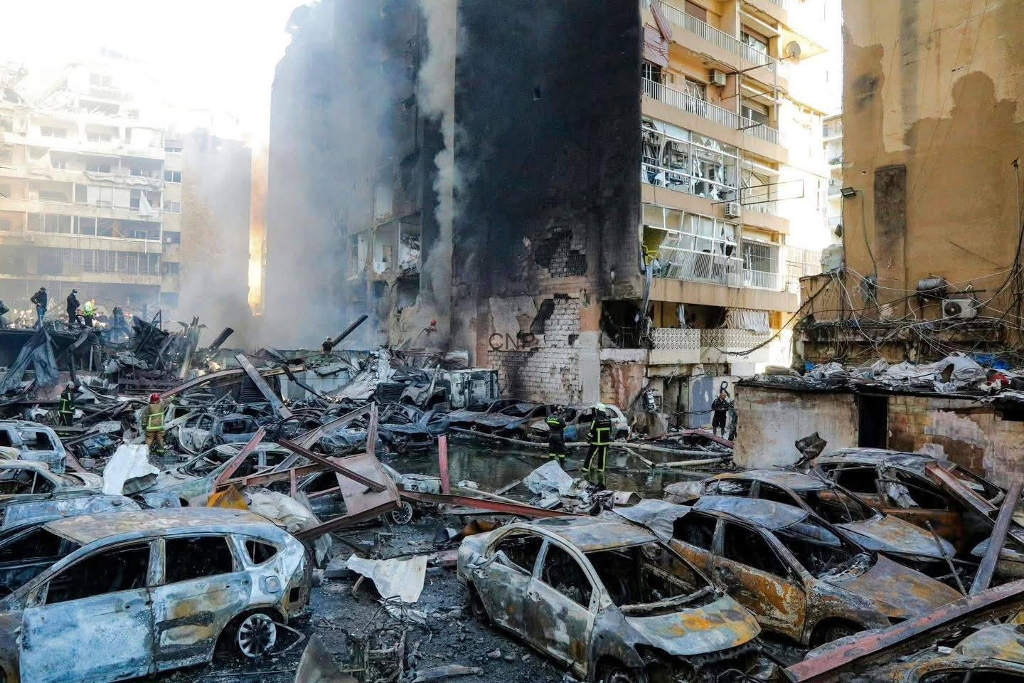 Firefighters amid charred ruins and collapsed cars in a destroyed urban block after a disaster.